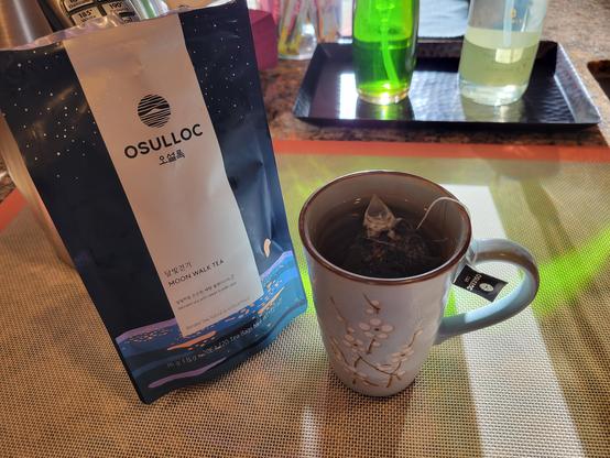 Photo of Osulloc tea bag and blue ceramic tea mug with brewing tea, on a kitchen counter with afternoon sunlight reflecting through a bottle of green dish soap liquid.