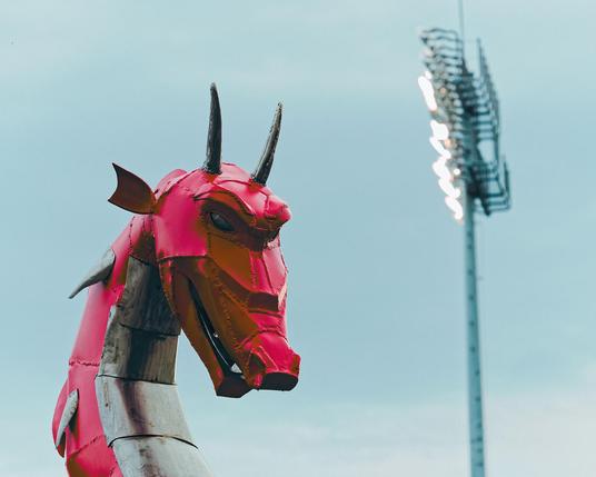 A small-to-medium metallic red dragon (Sorry Gygax, I see this one here) with horns and fierce eyes looks to the right from the left side of the image, on the right a set of stadium lights tower above him;  oddly if you ignored depth cues you could image he was being lit by the stadium lights;  behind those is an indistinct blueish-whiteish cloudy sky