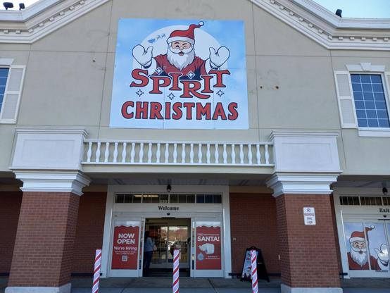 A picture of the outside of a Spirit Christmas store with a large sign with Santa on it. The bollards outside are candy can striped.