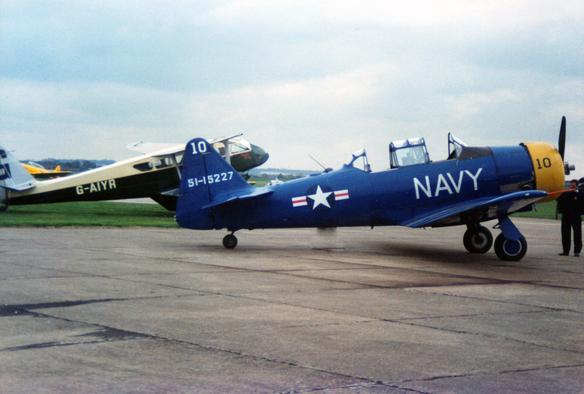 This image shows a blue Navy aircraft with the number 10 and the identification number 51-15227 parked on tarmac. The aircraft has a yellow nose and a white star insignia on its side. In the background, there is another aircraft with the registration G-AIYR. The focus is on the vintage military aircraft, highlighting their historical significance and design.