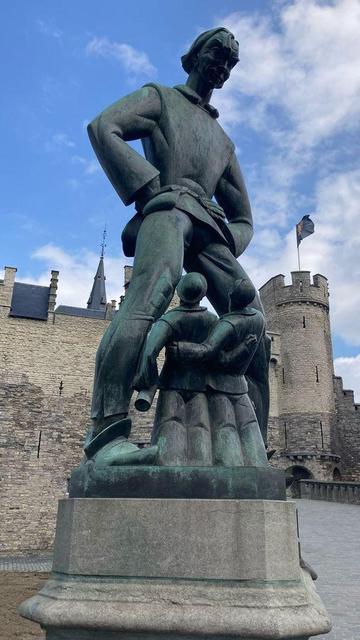 A large bronze statue of a tall man standing with his hands on his hips, looking down confidently. Below him, two smaller figures appear to be huddled together, almost in awe or fear of the large figure towering above them. The sculpture seems to convey a sense of dominance or protection by the larger figure. Behind the statue, there’s a stone building or castle with battlements and a tower, suggesting the statue is located in a historical setting. The sky above is partly cloudy.