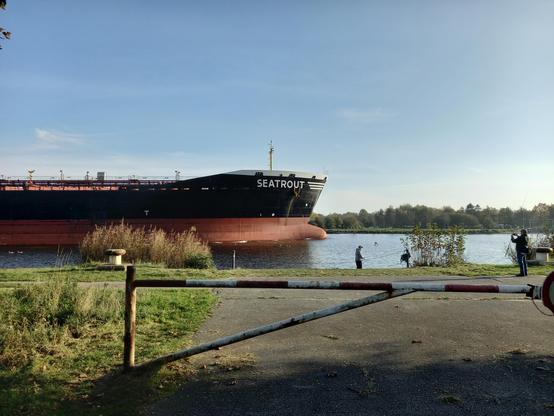 Auf dem Kanal kommt gerade ein ziemlich großes Frachtschiff mit Namen Seatrout angefahren und ist halb im Bild. Es ist sonnig und am Wasser steht ein Angler.