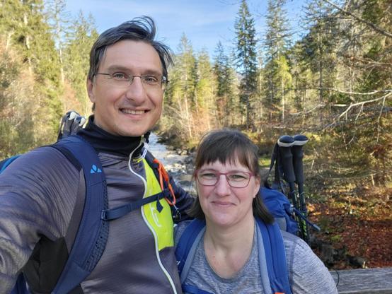 A man and a woman are seen taking a selfie outdoors. The man is wearing a backpack and glasses, while the woman has a backpack and trekking poles. They are both smiling in the selfie. The woman's neck and the backpack are shown in close-up shots. Additionally, there is a close-up of a body of water in the background. The man is wearing glasses and the woman is smiling. The scene depicts outdoor recreation, with a focus on hiking and backpacking. The dominant colors in the image are grey and blue, with a hint of green from the surrounding trees.
