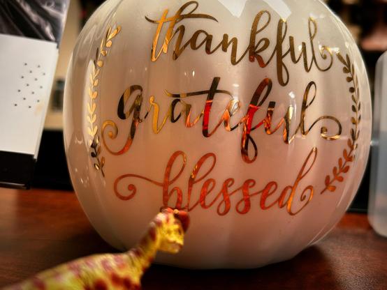 A green Dilophosaurus with brown spots stands before a glass pumpkin decoration that reads: “Thankful, Grateful, Blessed”