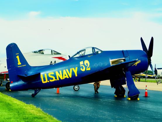 Alt text: A blue U.S. Navy aircraft with the number 52 and the text “U.S. NAVY” painted in yellow on its side is parked on an airstrip. The tail of the aircraft has the number 1 and the text “COMMANDER BLUE ANGELS” in yellow. The aircraft is a vintage propeller plane, and there is another aircraft in the background with the number 44 visible. The scene is set on a clear day with a blue sky.

