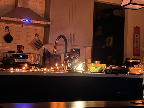 Ceramic baphomet sits behind a candle with a tall flame on a kitchen counter decorated with orange lights for Halloween.