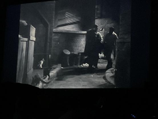 Three shadowy figures carrying a limp body into an alley filled with trash cans, and three cats in a black-and-white movie scene