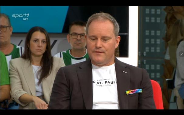 Bildbeschreibung von KI:
Das Bild zeigt eine Gesprächsszene im Studio von "Sport1 live". Im Vordergrund sitzt ein Mann, der einen dunklen Blazer und ein weißes T-Shirt mit der Aufschrift "ANTI-FASCIST FC ST. PAULI FOOTBALL CLUB" trägt. An seinem Blazer ist ein bunter Anstecker mit dem Schriftzug „Vielfalt“ zu sehen, der in Regenbogenfarben gestaltet ist und für Vielfalt und Toleranz steht. Der Mann scheint konzentriert oder nachdenklich zu sein und schaut leicht nach unten.

Hinter ihm sitzen drei Personen, die als Publikum wirken. Zwei von ihnen tragen grün-weiße Trikots, die möglicherweise auf eine Fußballmannschaft hinweisen. Die Frau links im Bild trägt einen cremefarbenen Blazer und schaut ebenfalls aufmerksam in die Richtung des Gesprächs.