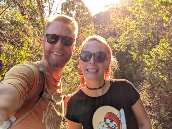 Photo of myself & my partner. My hair is pulled back, wearing sunglasses, facial piercings, black choker with a TeeFury shirt of Sally pinup style. My partner is wearing sunglasses, orange brewfest shirt & carrying a wooden walking stick. 10/21/2024.