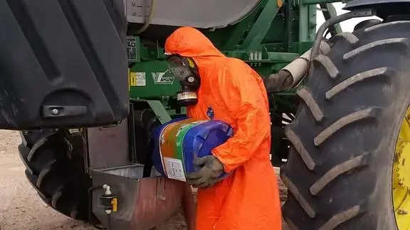 Wearing an orange all body & head orange suit, reinforced gloves and a safety breathing mask a toxic chemical herbicide liquid is poured into a tank on the side of a large tractor for spray dispersal onto fields.