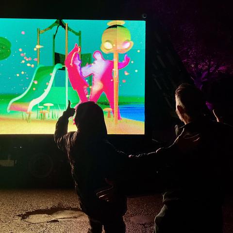 A brightly lit LED wall screen outside at night with a pair of silhouetted people in front. On the screen there is a colourful 3D rendition of a play park, with a slide in the background. In the foreground are two pink figures mirroring the pose of the two people. The pink figures are lit by virtual lights in the 3D scene shining from the top of a pole.