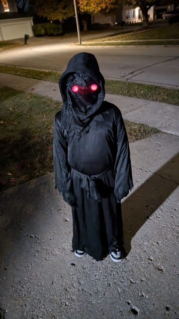 Small child standing next to a sidewalk dressed in a black robe, black hood, and black mask with glowing red eyes. 