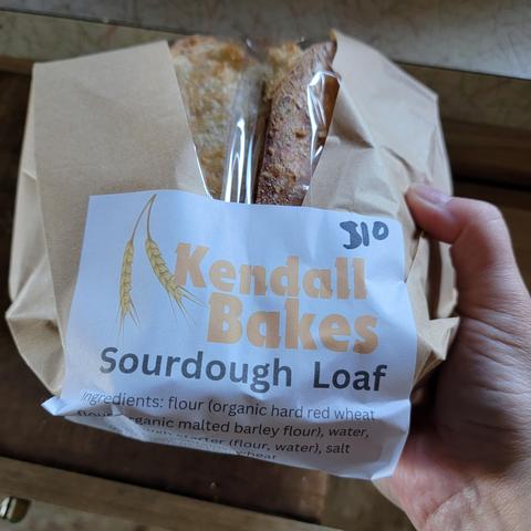 Fresh round loaf of homemade sourdough bread packaged in a brown paper bag labeled "Kendall Bakes."