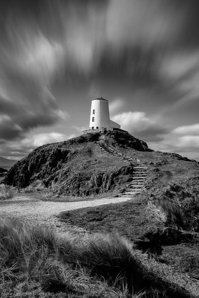 This monochrome long exposure photograph features Twr Mawr Lighthouse on Llanddwyn Island in Anglesey, North Wales. It captures the steps leading up to the historic lighthouse, which stands tall against a dramatic sky. The clouds streak across the image, conveying a sense of movement and adding depth to the scene. The composition highlights the sturdy structure of the lighthouse and the inviting path that guides viewers toward it. The piece is titled 'Tŵr Mawr Lighthouse.'