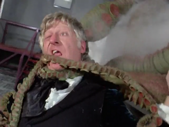 Jon Pertwee episode. The Doctor stands in a factory, in front of a machine, with large green tentacles and steam coming out of it. The tentacles wrap all around his body and neck, and he’s making a silly face with one eye closed and his mouth partially open. No caption.
