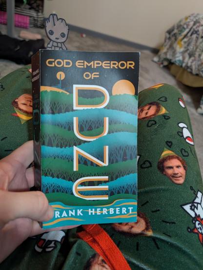 picture of me sitting in my reading chair. in view is the book God emperor of dune by Frank Herbert. I am wearing Will Ferrell elf pajama bottoms because it is cold and they're soft and fuzzy, also delightful. very similar to this book. I am surprised after all that has happened in this series that 3,500 years of the god emperor's rule can bring about a new story. what a Craftsman Frank Herbert was.