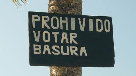 Attached to trunk of a tree is a sign with hand-painted letters reading Prohivido Votar Basura. It should read Prohibido Botar Basura