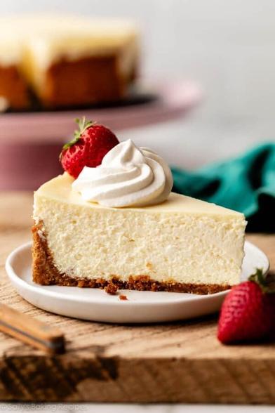An image of a very fluffy slice of cheesecake on a plate, topped with a fresh strawberry and whipped cream.