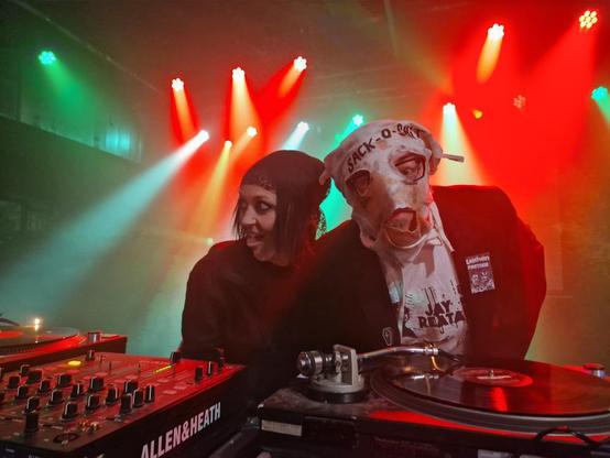 Zwei verkleidete Personen an einem DJ-Pult auf einer Bühne mit farbigen Scheinwerfern im Hintergrund.