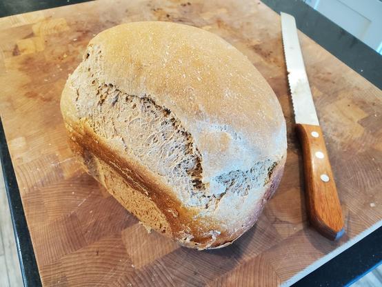 A small loaf of brown bread on a cutting board. A bread knife lies next to it. The smooth top has a natural tear in it probably from yeast activity.