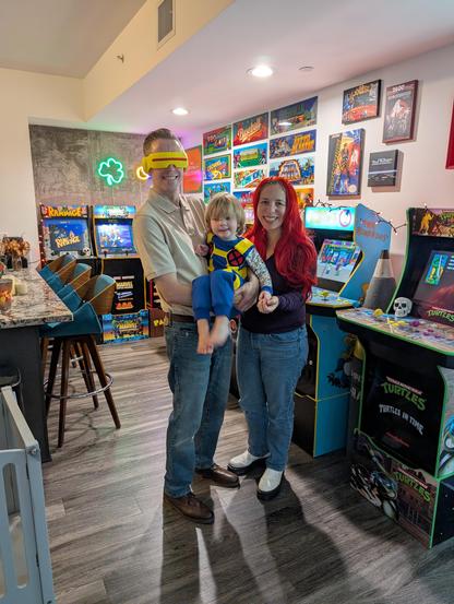 Patrick, Kara, and PJ dressed for Halloween as Cyclops from X-Men '97, Jean Grey from X-Men '97, and Cable from X-Men, respectively. They are standing in front of arcade machines.