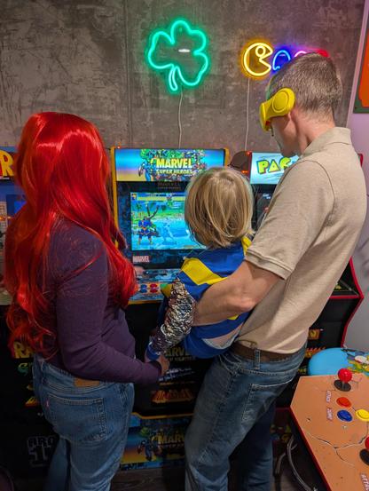 Patrick, Kara, and PJ dressed for Halloween as Cyclops from X-Men '97, Jean Grey from X-Men '97, and Cable from X-Men, respectively. They are facing away from the camera, looking at a Marvel Super Heroes arcade machine. Cyclops is on the screen.