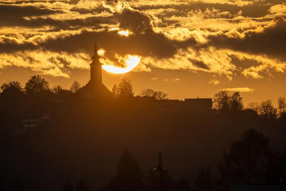 Aufnahme mit einem 600er-Teleobjektiv, die Kirche von Frauenberg und der Hügel auf dem sie steht als Silhouette. Die Sonnenkugel scheint den Kirchturm zu streifen.  