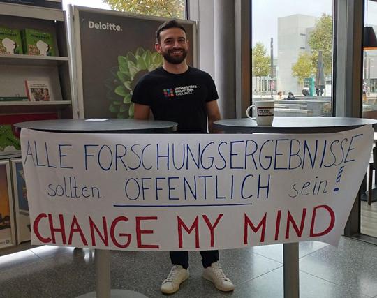 Deutsch: Ein Mitarbeiter der Universitätsbibliothek steht hinter einem Stand auf der Open Access Week der TU Chemnitz und auf einem Banner im Vordergrund zu sehen steht der Text "Alle Forschungsergebnisse sollten öffentlich sein! Change my Mind"

English: An employee of the university library stands behind a stand at Chemnitz University of Technology's Open Access Week and a banner in the foreground reads “All research results should be public! Change my Mind”