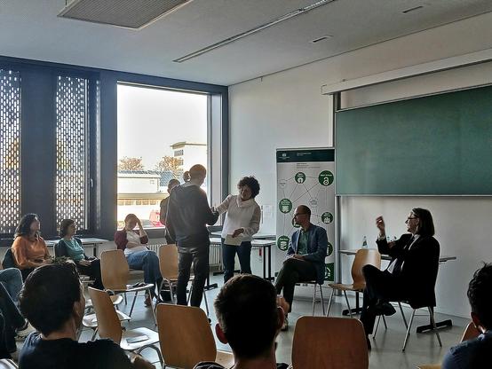 Deutsch: Im Rahmen der Podiumsdiskussion überreicht Angela Malz (Direktorin der Universitätsbibliothek) den Open Access Award an einen Preisträger. Im Vordergrund sitzen in kreisförmig aufgestellten Stuhlreihen weitere Teilnehmer.

English: During the panel discussion, Angela Malz (Director of the University Library) presents the Open Access Award to an award winner. In the foreground, other participants sit in rows of chairs arranged in a circle. 