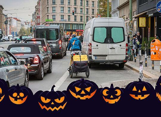 Straße mit vielen Autos, Fahrradfahrer schlängeln sich durch den starken Verkehr, da Radwege zugeparkt sind. Im Vordergrund grinsende Halloween-Kürbise.