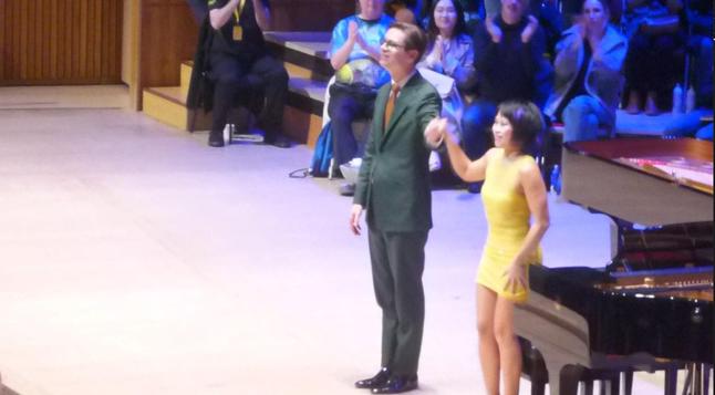 Yuja Wang and Víkingur Ólafsson on stage taking a bow. Piano and audience members behind them. Yuja in mini-skirted yellow dress, Vikingur in Lincoln green suit with rust tie and white shirt.