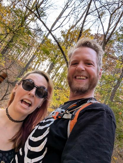 Selfie photo of myself & my partner with the autumn trees behind us. I'm wearing a black skull tank top, sunglasses, facial piercings. My partner is wearing a black skull t-shirt, orange backpack & holding a walking stick. 