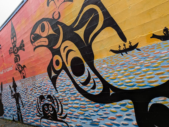 The continuation of the mural still has a background of vast water, but the sky is in warm orange and yellow tones, not cool blue, and the hills are gone. A large fish emerges from the water and faces the walking humanoid approaching it. A crab, flying bird, and another jumping fish seem to witness the scene. People in canoes with hats woven from grass or other plant material travel in canoes, approaching the center. One of them stands at the shore, gesturing for the others to make land or signaling his intention to speak.