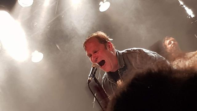Bildbeschreibung von KI:
Das Foto zeigt eine energetische Live-Performance auf einer Bühne. Im Vordergrund sieht man einen Mann, der intensiv in ein Mikrofon singt oder schreit. Er hat kurzes, leicht feuchtes Haar und trägt eine Jeansjacke. Die Beleuchtung erzeugt einen dramatischen Effekt mit starken Lichtstrahlen und Nebel, was der Szene eine dynamische und emotionale Stimmung verleiht. Im Hintergrund ist ein weiterer Mann zu erkennen, der möglicherweise ein Instrument spielt. Die Atmosphäre wirkt leidenschaftlich und intensiv, als ob die Künstler tief in ihre Musik vertieft sind. 