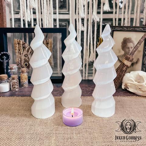 Three specimens of the Selenite Unicorn Horn are standing on a burlap-covered surface. They are a shimmery translucent white and are carved into tall spiral shapes that end in a point. Presented by Inked Goddess Creations.