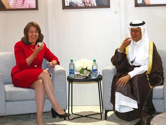 Alberta premiere Danielle Smith in a red dress and a man in traditional Middle Eastern attire sit together on a couch, both smiling and holding up cups. Water bottles and a flower arrangement are on a nearby table. Photographs hang on the wall behind them.