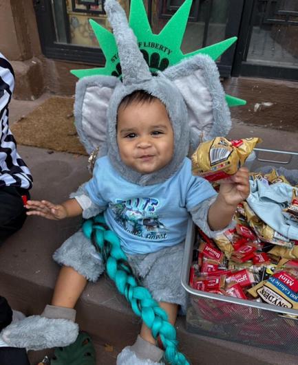 An infant sitting on a curb in some NY Liberty gear and also an Ellie the Elephant costume. The child is really cute and holding up a small bag of snacks for the camera.