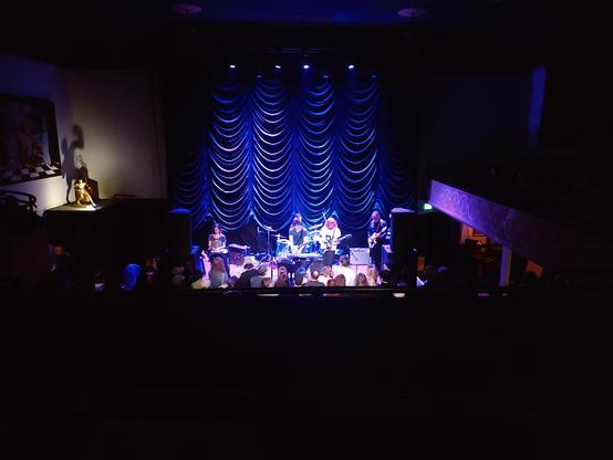 The band Swirlies on stage at the Mission Theater in Portland Oregon. Photo taken from the balcony so it's a fairly wide shot of the whole venue with the band lit in blue. Some of the crowd is visible and some ripply curtains behind the stage are also blue/purple.