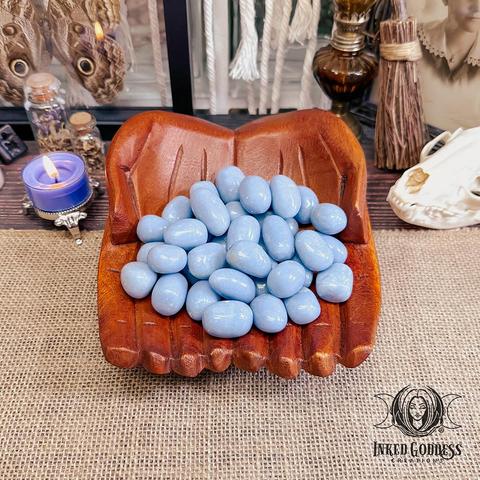 A variety of Angelite Tumbled Gemstone crystals rest in cupped, carved wooden hands. The crystals are roughly spherical and are a glossy sky blue with small white and beige inclusions. Presented by Inked Goddess Creations.