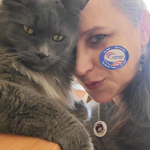 A fluffy dark grey cat leans against a Caucasian woman with an "I Voted" sticker on her left cheek.