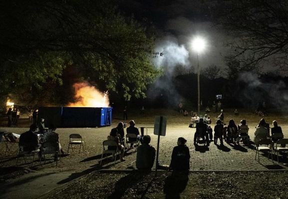 A night scene featuring a group of people sitting on chairs, watching smoke and flames rising from a blue dumpster. Surrounding trees and ambient lighting contribute to the atmosphere.