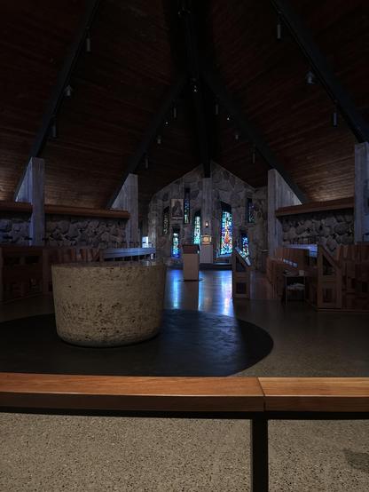 An interior view of a chapel featuring a stone altar in the foreground, wooden benches, and large stained glass windows illuminating the space. The ceiling is high and wooden, creating a serene atmosphere.