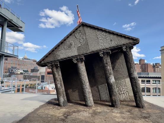 Photo of a large outside art installation at the Whitney Museum. The piece shows the White House covered with dirt and tilting over, ready to fall like the Leaning Tower of Pisa. The American flag on top is upside down.