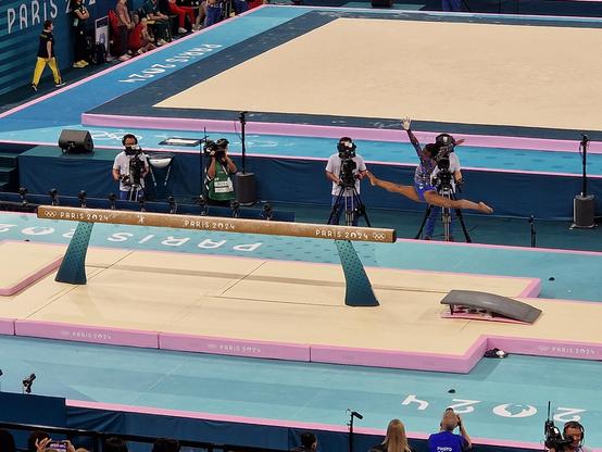 Simone Biles in perfecte spagaat in vlucht tijdens de opsprong op de evenwichtsbalk. Rechterbeen voor, armen hoog en opzij gestoken.  