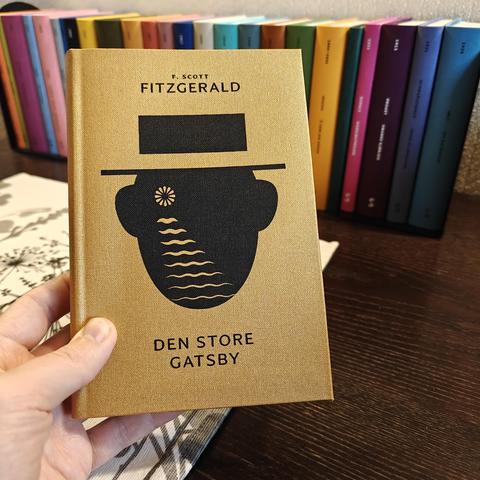 Eg held opp boka Den store Gatsby av F. Scott Fitzgerald. Omslaget er gullfarga og er illustrert med ein figur som ser ut som eit hovud med hatt. Vi ser ingen detaljar i det einsfarga fjeset, men der det eine auget skulle ha vore, ser vi ein strålande sirkel - kanskje ei lykt eller månen på horisonten? Under sirkelen er det ei rad med bølgjer. Bølgjene blir lenger di lenger ned på fjeset dei kjem, noko som illuderer til at sirkelen er eit lyspunkt som vi ser på avstand over vatn. Dette gir òg assosiasjonar til tårar. 
I bakgrunnen står alle dei andre bøkene i nynorske klassikarar-serien til Skald forlag.