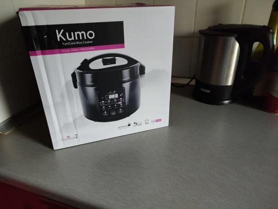 An image showing the package of a Kumo rice cooker from Yum Asia in black on a grey countertop. The rice cooker looks like the old traditional rice cookers but with an electric panel for control instead of the cook and keep warm switch.