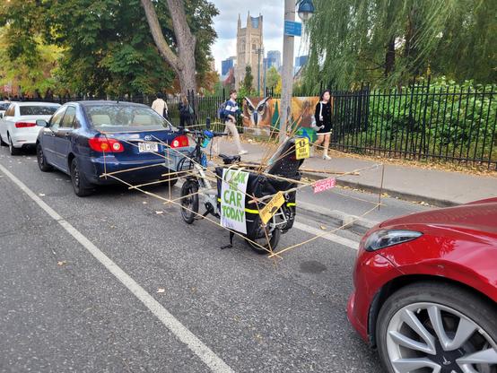 My cargo bike parked in a street parking spot with a bamboo frame the size of a car built around it and a sign on the side that says "I TOOK THE CAR TODAY"