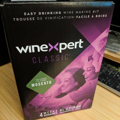 A square cropped photo of a wine making kit. It says winexpert classic, califonia  moscato, easy drinking wine making kit trousse de vinification - facile a boire oui oui... The box is back and pink and has happy people on it. The people are in fact people, humans if you will.. it says some stuff at the bottom but the preview is blocking that and I'm in a lazy mood to alt tab and look.. it's just numbers or more french.