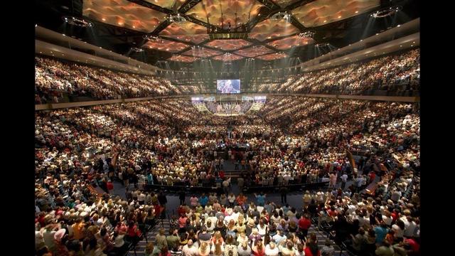 A large crowd of people gathered in an arena, in a megachurch event. The venue is brightly lit, showcasing a vibrant atmosphere with many seated attendees.