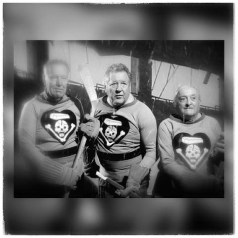 A black-and-white film still of three old white men in strange hockey uniforms, while holding hockey sticks and wearing oversized, thick hockey gloves. The logo on the front of their jerseys includes a skull and two angled bones set atop a darkly colored heart.
